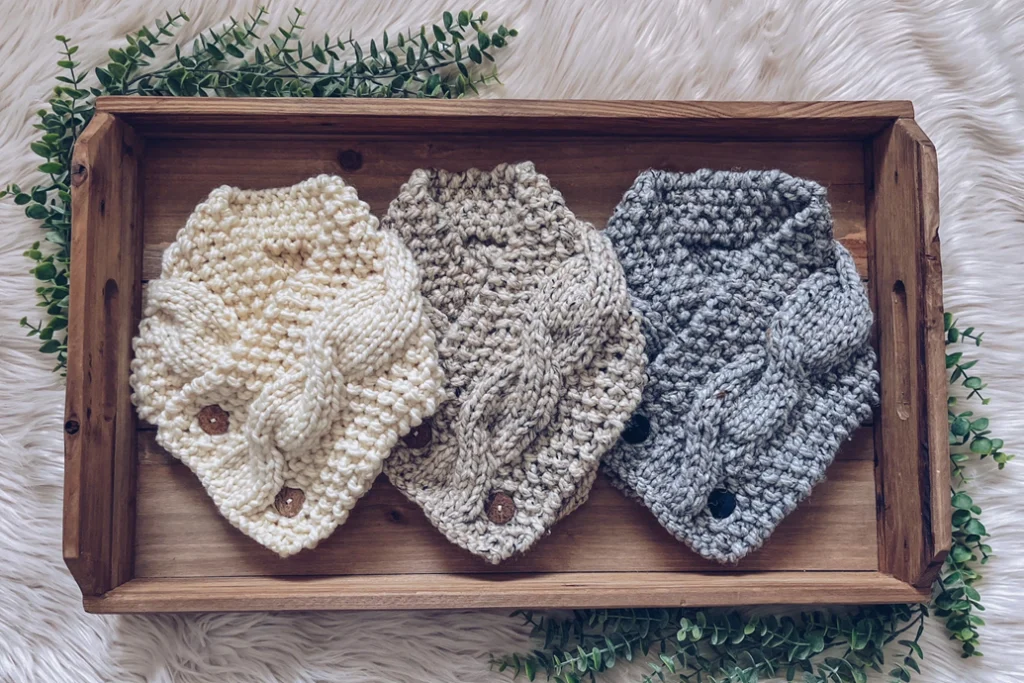 Three handmade knitted scarves in cream, beige, and gray on a wooden tray.