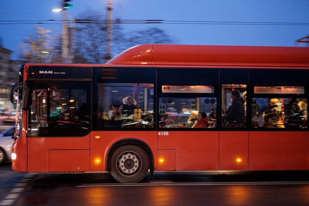 Red public bus carrying passengers through Vilnius at dusk.
